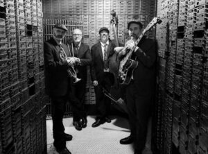 black and white photo of four men holding different instruments and standing in a bank vault